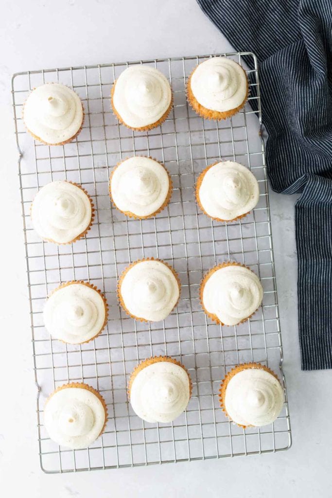 Twelve cupcakes with white frosting arranged on a wire cooling rack, next to a dark striped cloth on a light surface.