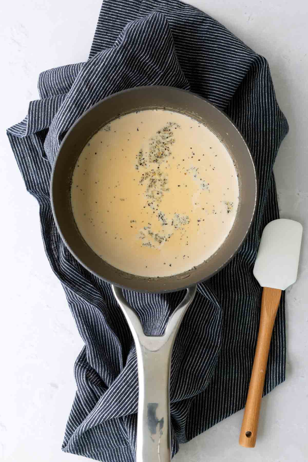A saucepan filled with a creamy sauce and specks of seasoning sits on a striped cloth next to a white spatula with a wooden handle.