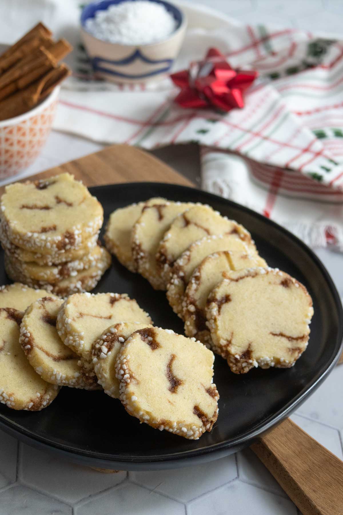 A black plate with several slices of sugar-dusted cookies, featuring swirls of filling, sits on a wooden board with holiday-themed items in the background.
