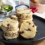 Stacks of round, sugar-coated cookies with brown swirls are arranged on a black plate atop a wooden board; holiday decorations and a bowl are in the background.