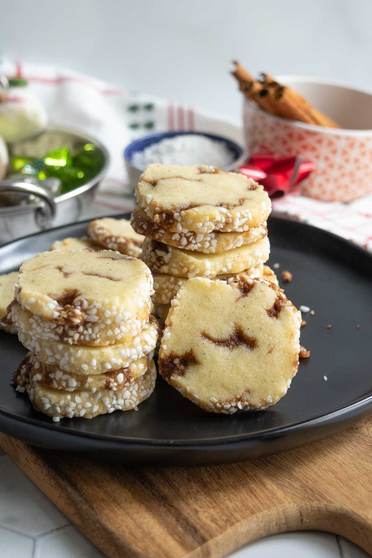 Stacks of round, sugar-coated cookies with brown swirls are arranged on a black plate atop a wooden board; holiday decorations and a bowl are in the background.