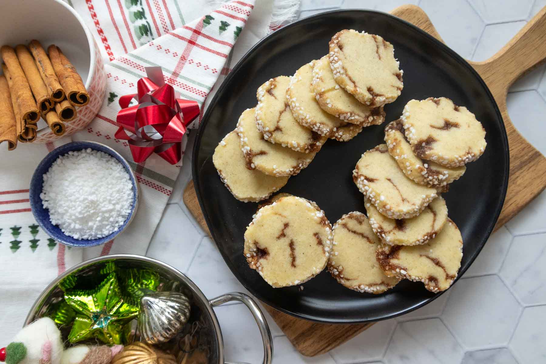 A black plate with stacks of round sugar cookies, some swirled with brown filling, sits on a wooden board next to baking ingredients and holiday decorations.