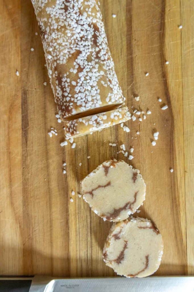 A roll of marbled cookie dough topped with coarse sugar on a wooden cutting board, with two sliced cookies and a knife nearby.