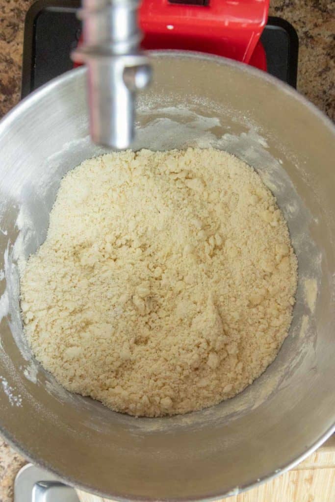 A metal mixing bowl with coarse, crumbly dough inside, attached to a stand mixer on a kitchen countertop.