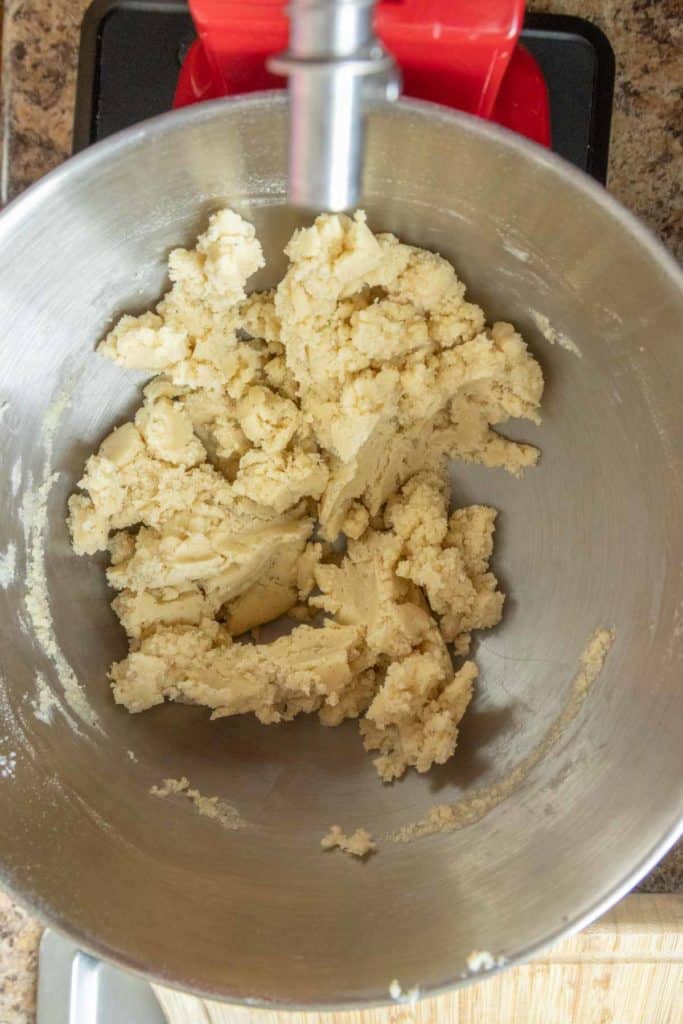 A metal mixing bowl containing a pale, crumbly dough mixture sits attached to a red stand mixer on a kitchen countertop.