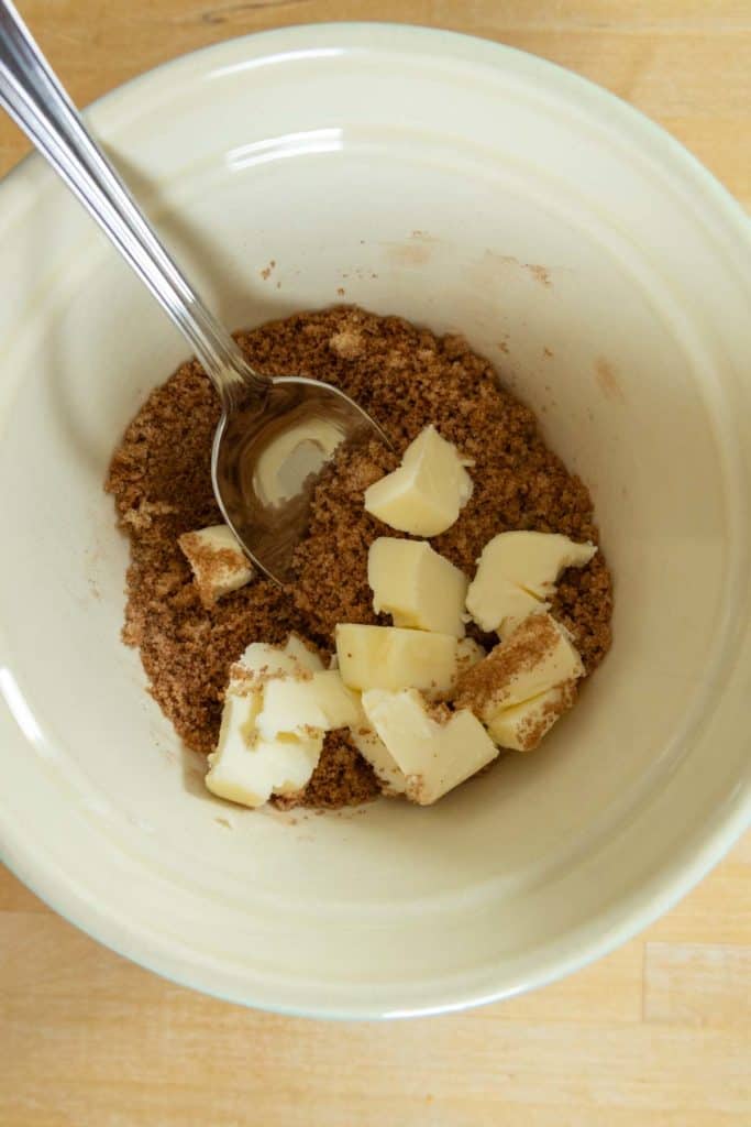 A mixing bowl containing brown sugar, cubed butter, and a metal spoon on a wooden surface.