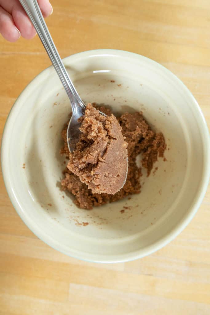 A spoon holds a portion of brown sugar paste inside a beige ceramic bowl on a light wooden surface.