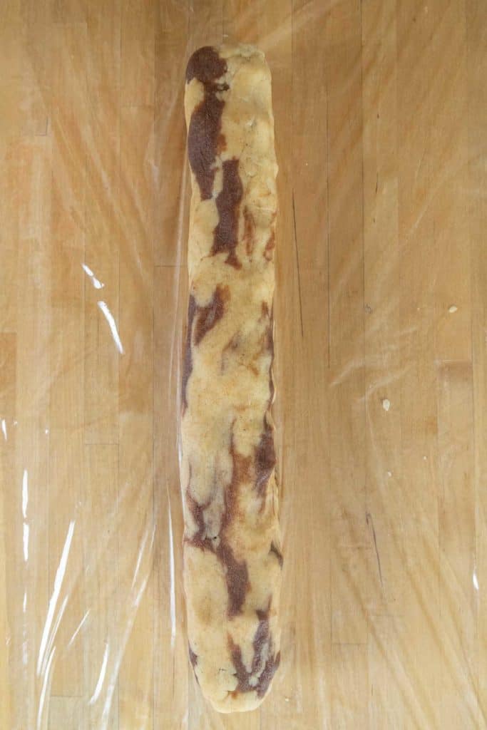 A log of marbled cookie dough rests on a sheet of plastic wrap on a wooden surface.