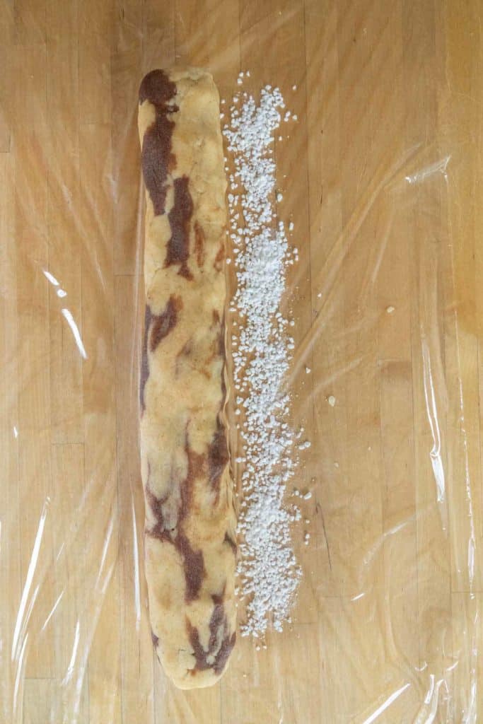 A rolled marbled dough log sits on plastic wrap next to a line of coarse white sugar on a wooden surface.