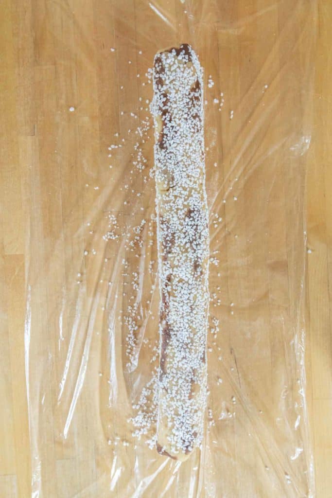 A long strip of dough rolled with filling and sprinkled with pearl sugar, resting on clear plastic wrap on a wooden surface.