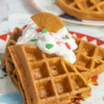Two golden brown waffles topped with whipped cream, festive red and green sprinkles, and a cookie piece, served on a decorative plate.