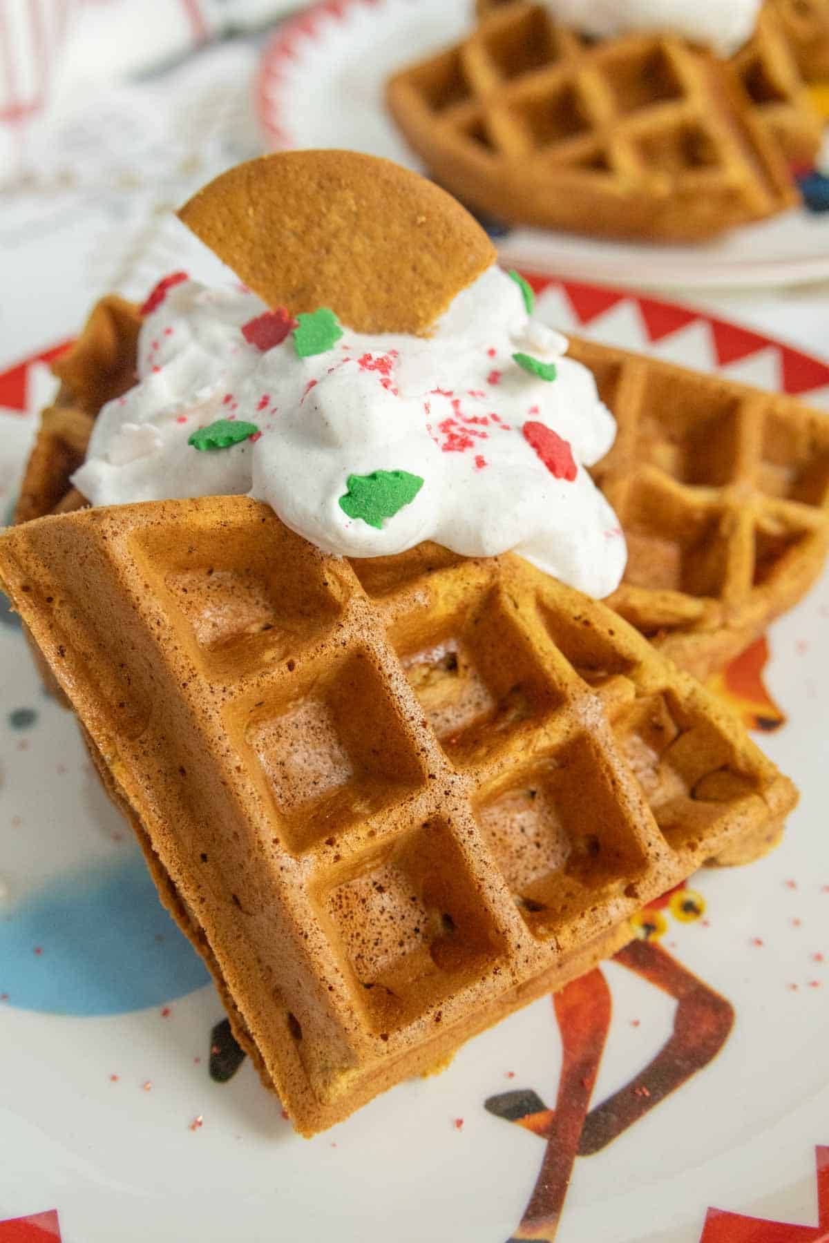 Gingerbread Waffles - If you need an idea for Christmas morning breakfast, why not give these Gingerbread Waffles a try? A plate with two thick waffles topped with whipped cream, red and green sprinkles, and a small round cookie piece.