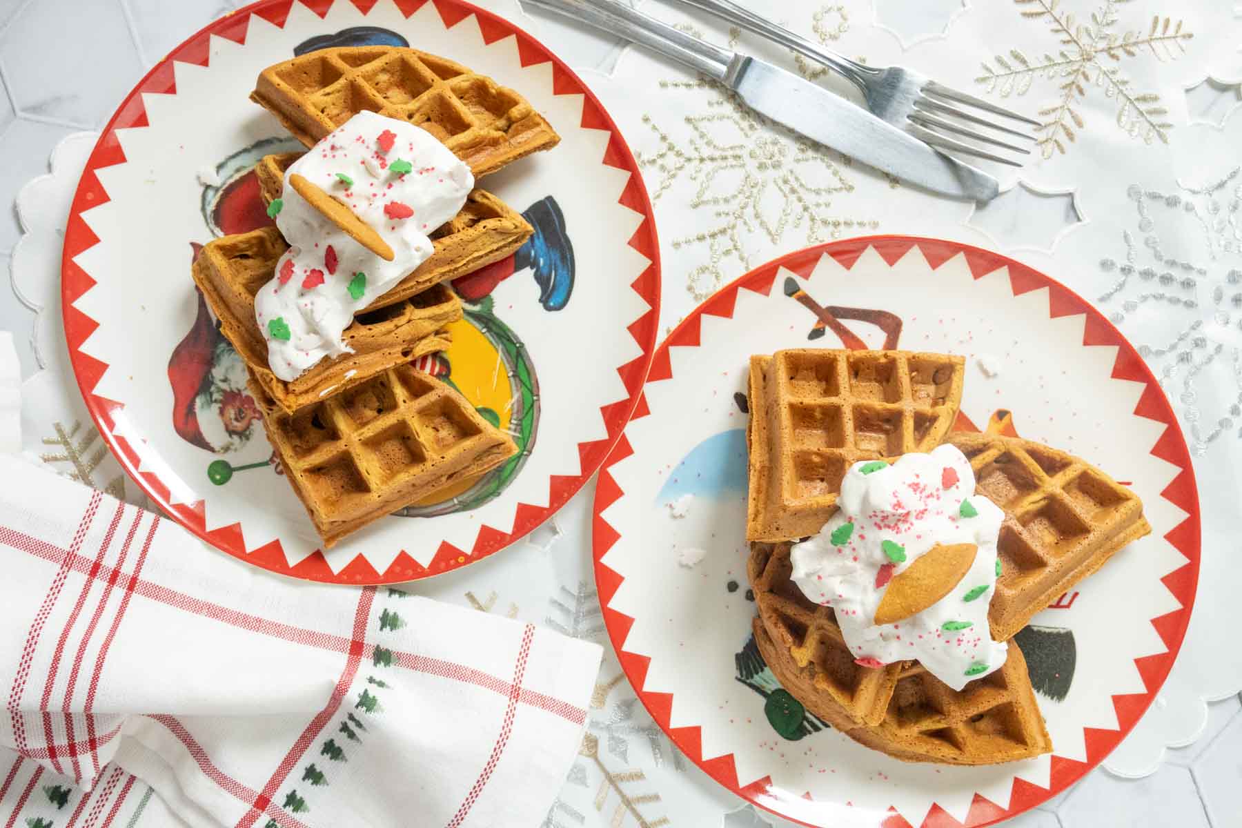 Two festive plates of waffles topped with whipped cream and sprinkles are set on a white tablecloth, with a fork, knife, and a red-and-white checkered napkin nearby.