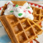 Close-up of gingerbread waffles topped with whipped cream and festive sprinkles on a decorative plate.