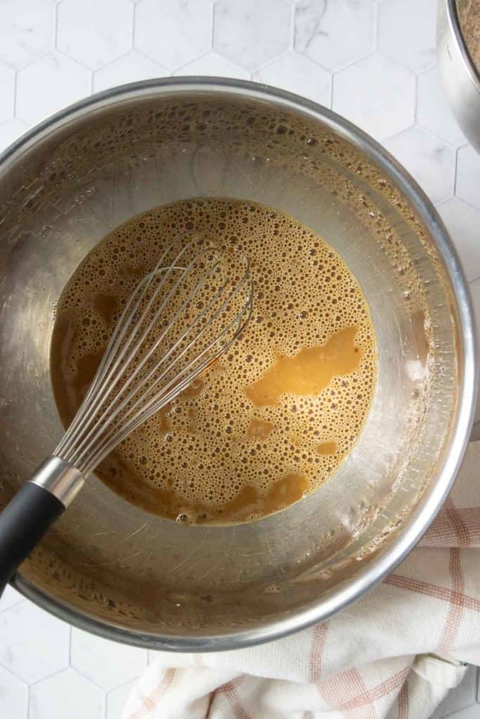 A metal mixing bowl contains a light brown liquid mixture with bubbles, and a whisk rests inside the bowl on a countertop.