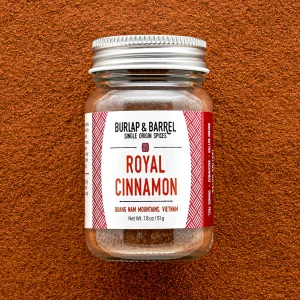 A clear glass jar labeled "Royal Cinnamon" from Burlap & Barrel sits on a brown textured surface, containing 1.8 oz of ground cinnamon sourced from the Quang Nam Mountains, Vietnam.