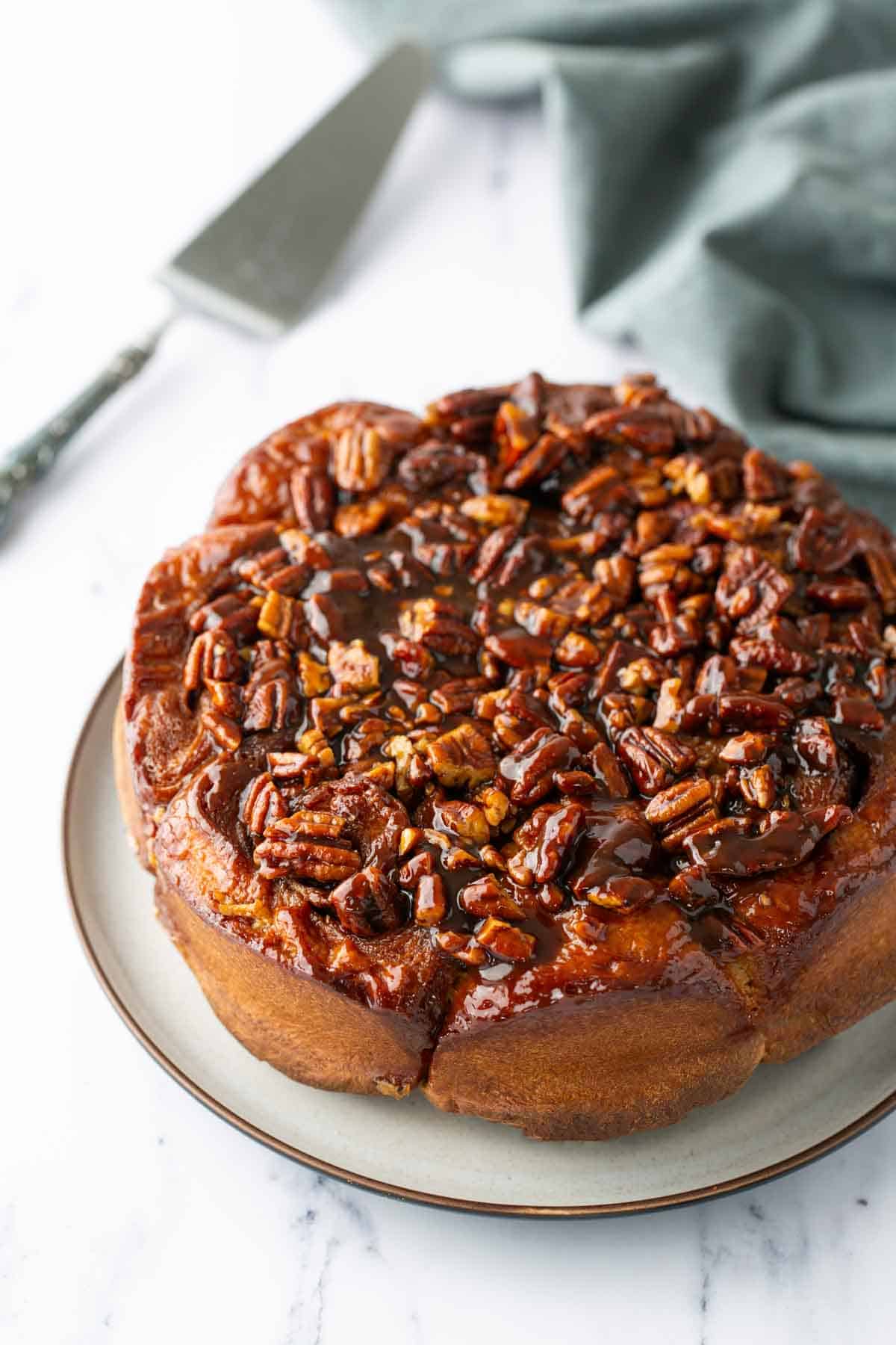 A round sticky bun topped with glazed pecans sits on a plate, with a cake server and a cloth in the background.