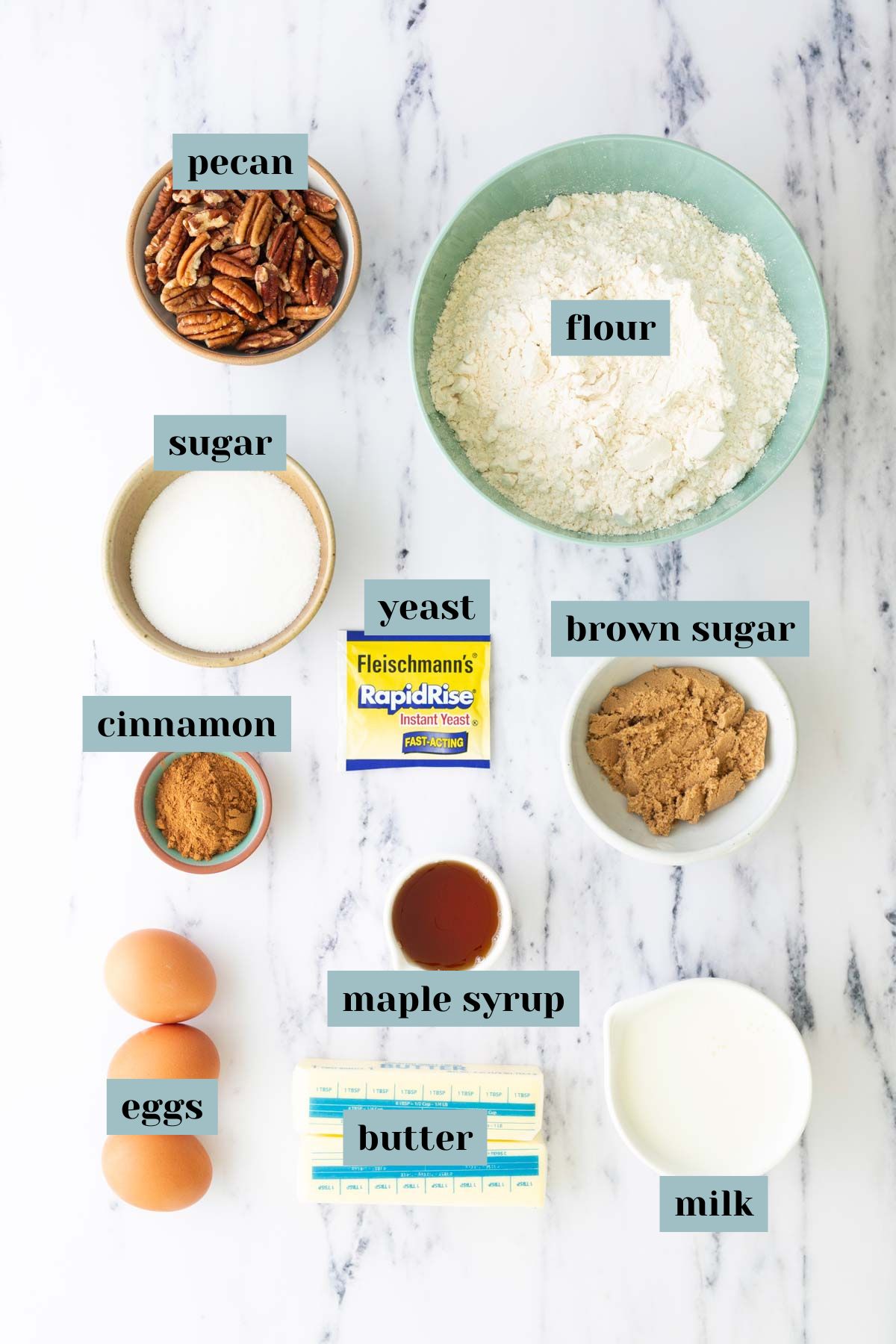 Baking ingredients on a marble surface, including pecans, flour, sugar, brown sugar, cinnamon, eggs, butter, milk, maple syrup, and a packet of rapid rise yeast.