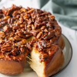 A round sticky bun topped with glazed pecans sits on a plate, with one piece cut and pulled slightly away, revealing its soft interior.