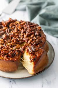A round sticky bun topped with glazed pecans sits on a plate, with one piece cut and pulled slightly away, revealing its soft interior.