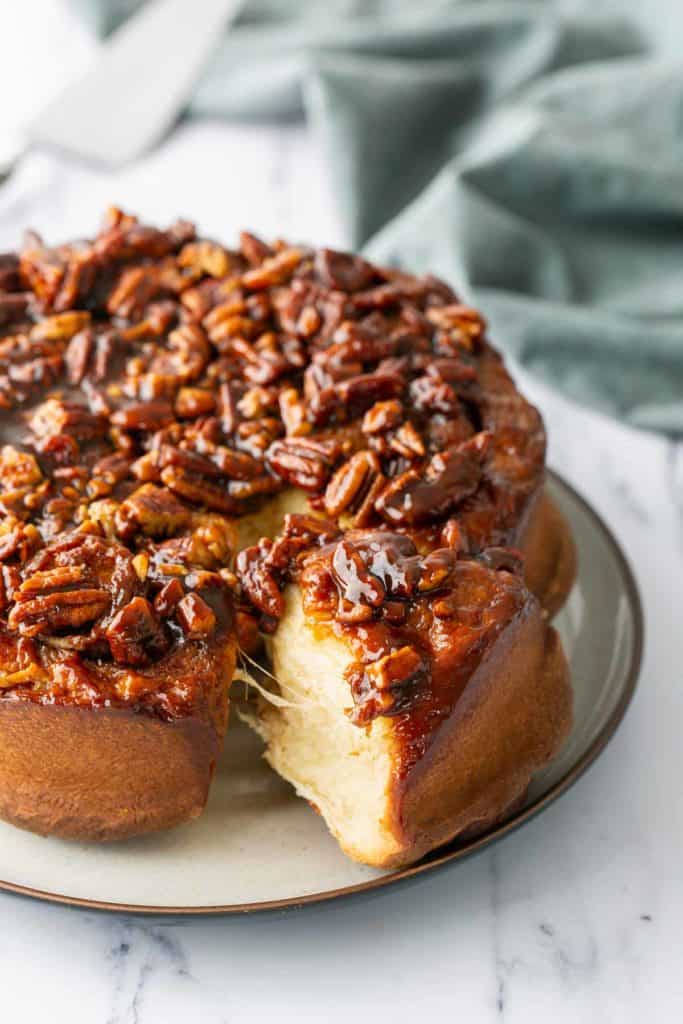 Sticky Buns A round sticky bun topped with glazed pecans sits on a plate, with one piece cut and pulled slightly away, revealing its soft interior.