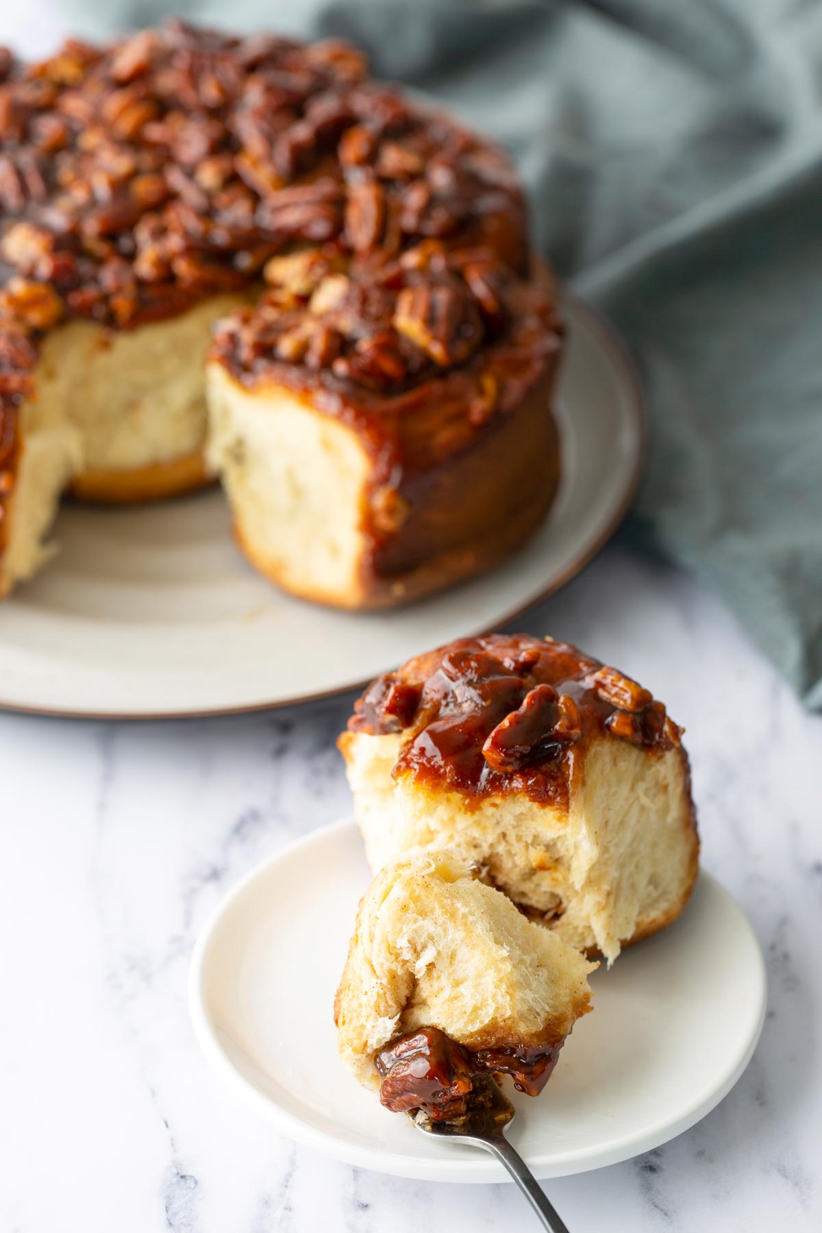 A sticky pecan bun served on a small white plate with a forkful cut out, with the rest of the buns on a plate in the background.