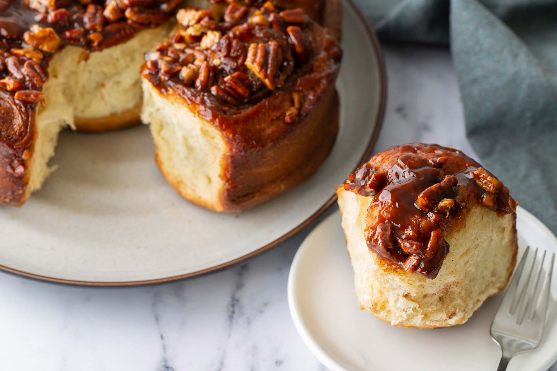 A sticky bun topped with pecans sits on a small plate with a fork, next to a larger plate holding more sticky buns.