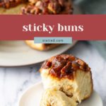 A sticky bun topped with caramelized pecans sits on a white plate, with a larger serving of sticky buns in the background.