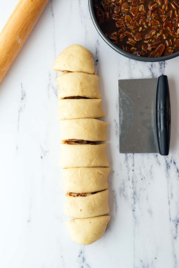 Sliced roll of dough filled with pecan mixture on a marble surface, next to a dough scraper, rolling pin, and bowl of pecan filling.