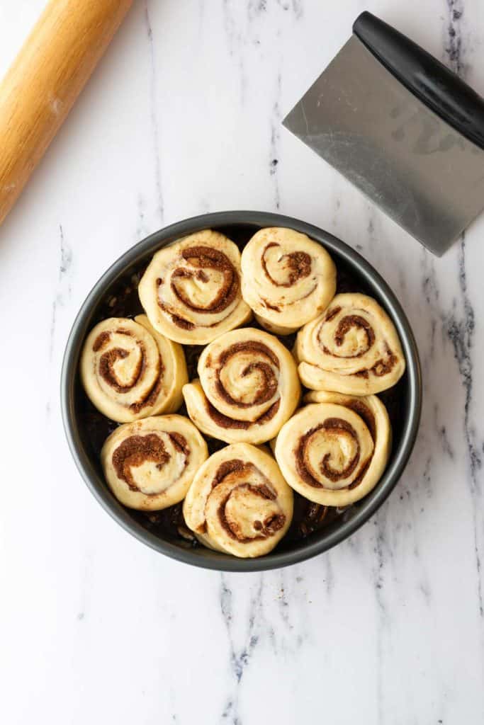 A round baking pan filled with unbaked cinnamon rolls sits on a marble surface next to a rolling pin and a dough scraper.