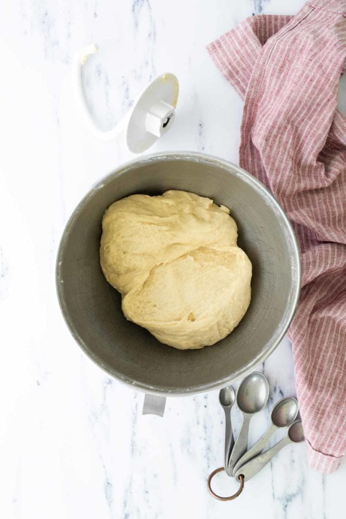 A metal mixing bowl with risen dough, surrounded by a red-striped cloth, a set of measuring spoons, and a stand mixer attachment on a white surface.