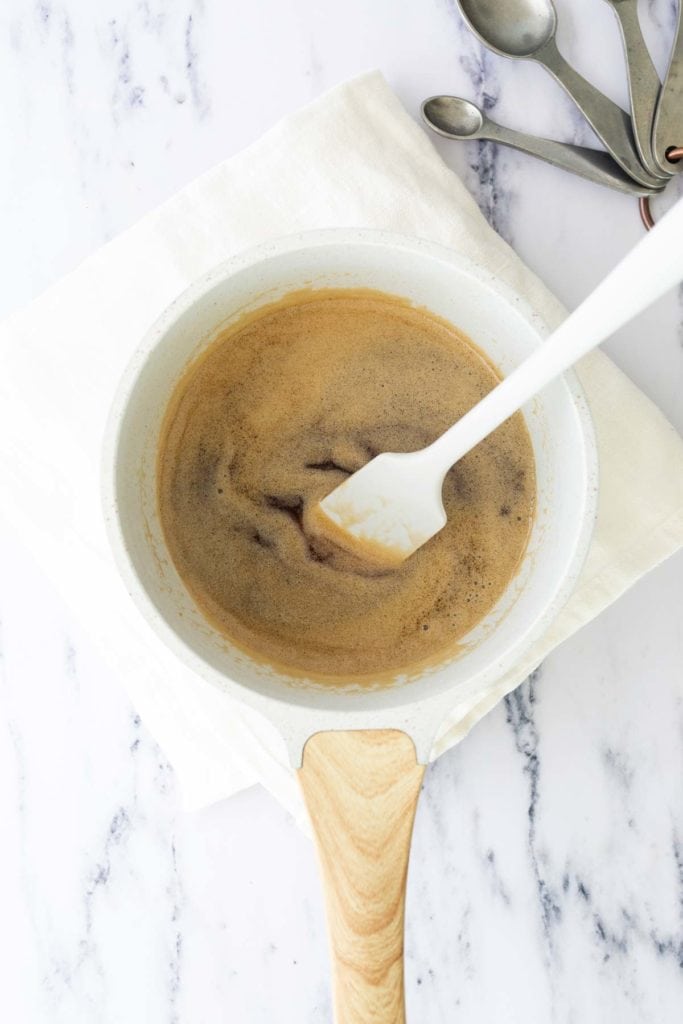 A white saucepan with a light wooden handle containing a brown liquid being stirred with a white spatula, placed on a white cloth next to metal measuring spoons.