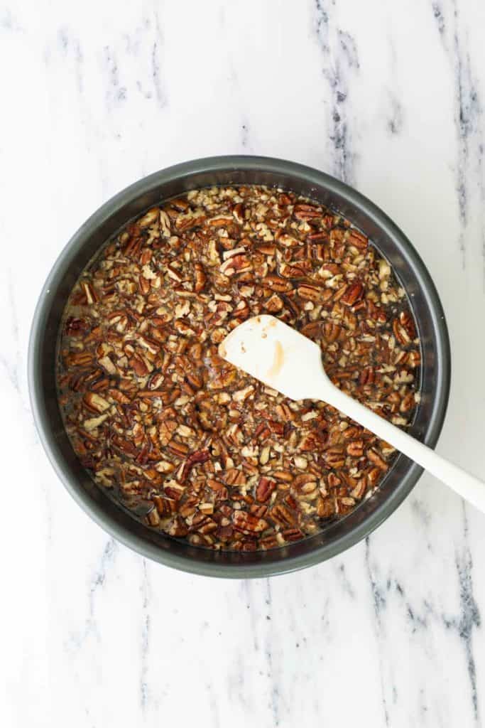Round baking pan filled with chopped pecans and batter, with a white spatula resting inside, on a white marble surface.