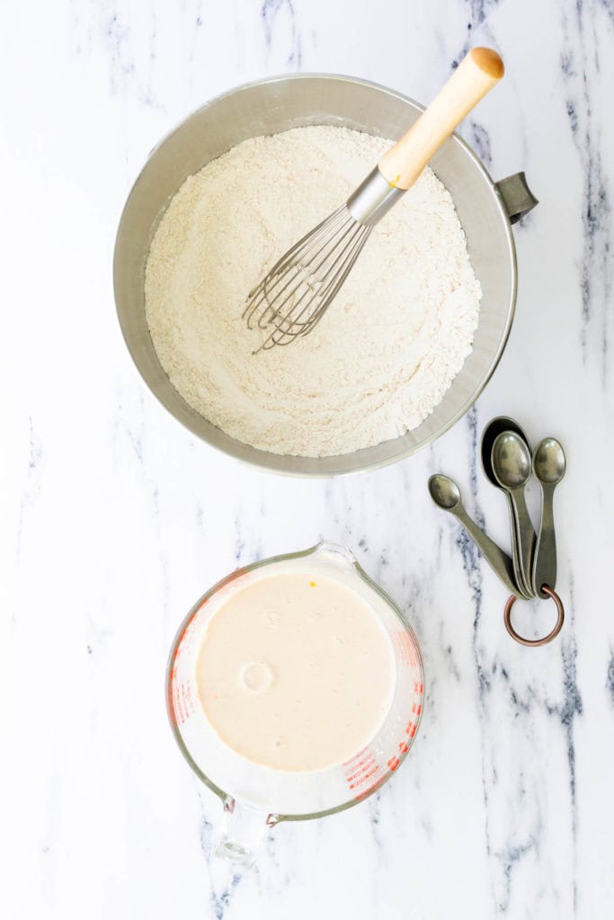 A metal bowl with flour and a whisk, a glass measuring cup with liquid batter, and a set of measuring spoons on a white marble surface.