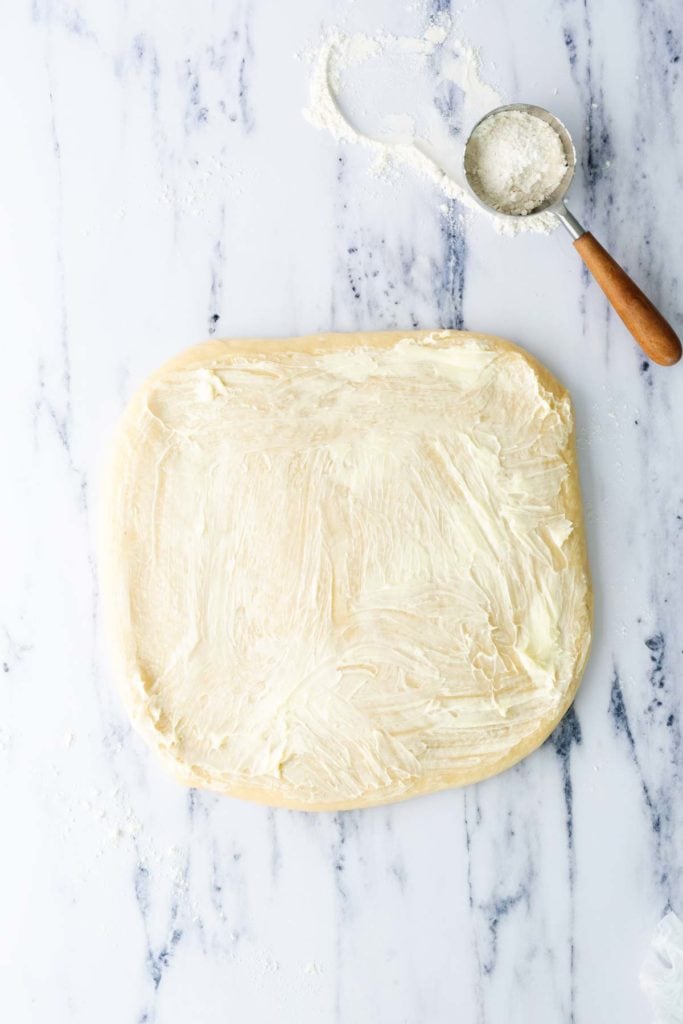 A square piece of dough is spread with butter on a marble surface next to a metal measuring cup with flour.
