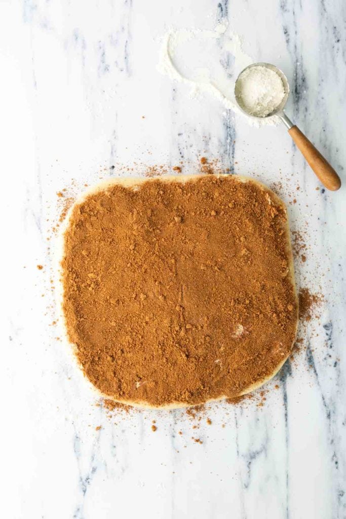 A rolled-out dough sheet covered with a layer of cinnamon-sugar mixture sits on a marble countertop next to a metal sifter with flour.
