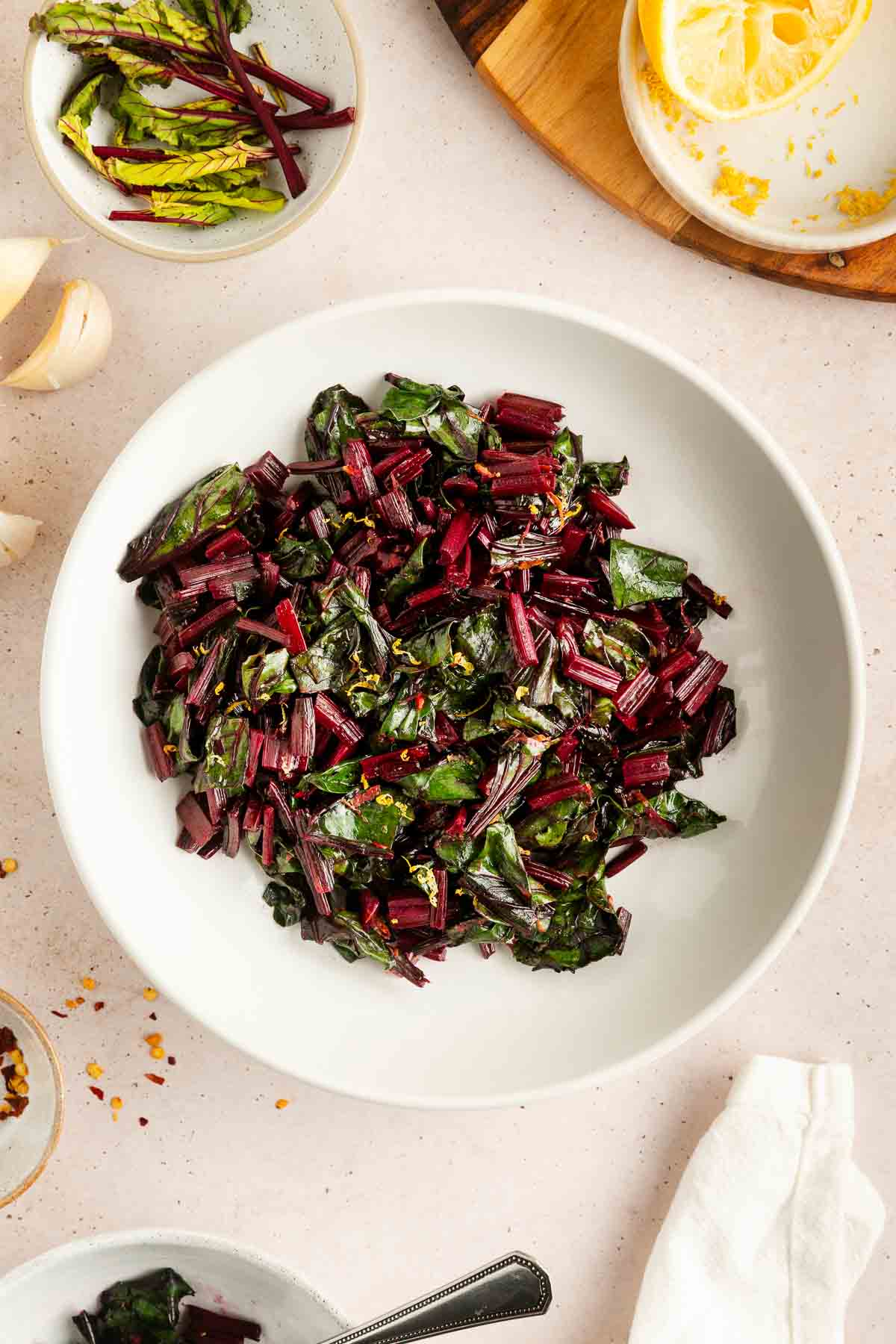 A white bowl filled with cooked beet greens and stems, surrounded by lemon, garlic, and additional beet greens on a light countertop.