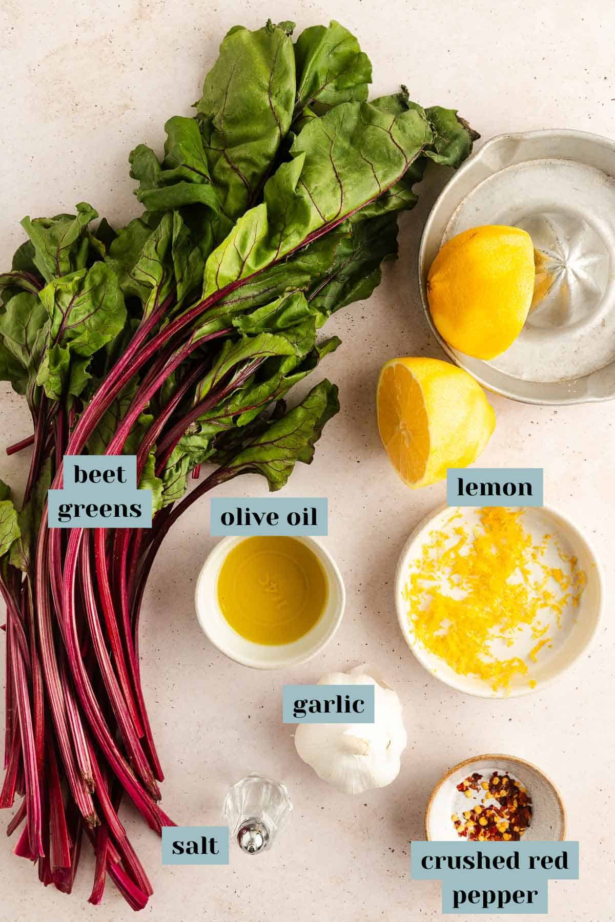 A bunch of beet greens, a halved lemon, a bowl of olive oil, minced garlic, salt, lemon zest, and crushed red pepper arranged on a light countertop.