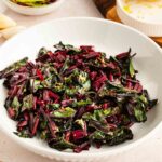 A white bowl filled with sautéed beet greens, garnished with lemon zest, sits on a table next to half a squeezed lemon and small dishes.