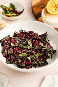 A white bowl filled with sautéed beet greens, garnished with lemon zest, sits on a table next to half a squeezed lemon and small dishes.