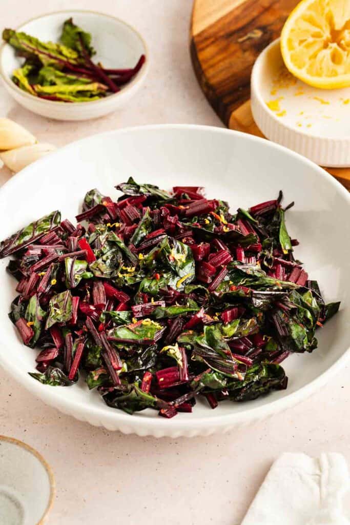 A white bowl filled with sautéed beet greens, garnished with lemon zest, sits on a table next to half a squeezed lemon and small dishes.