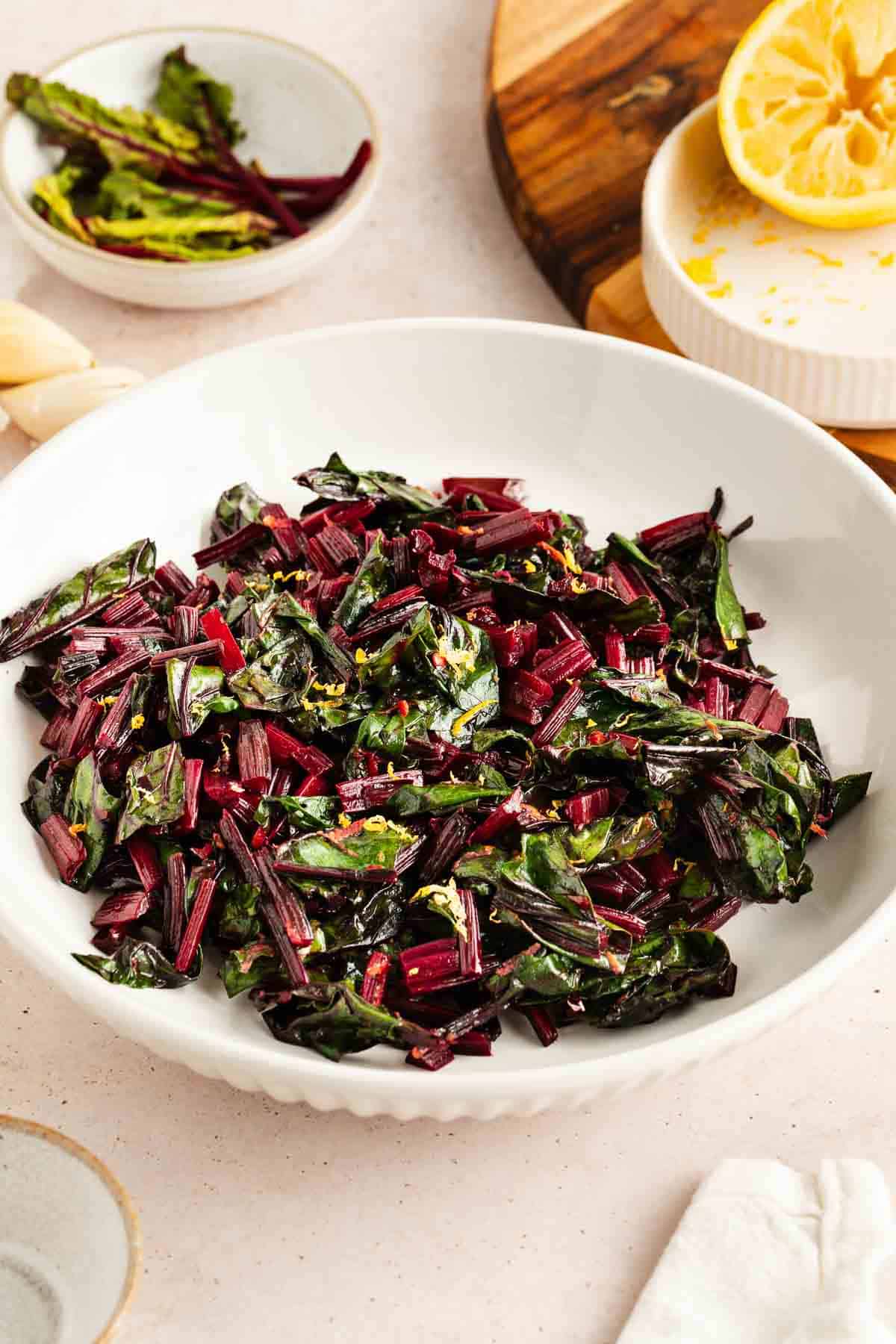 A white bowl filled with sautéed beet greens, garnished with lemon zest, sits on a table next to half a squeezed lemon and small dishes.