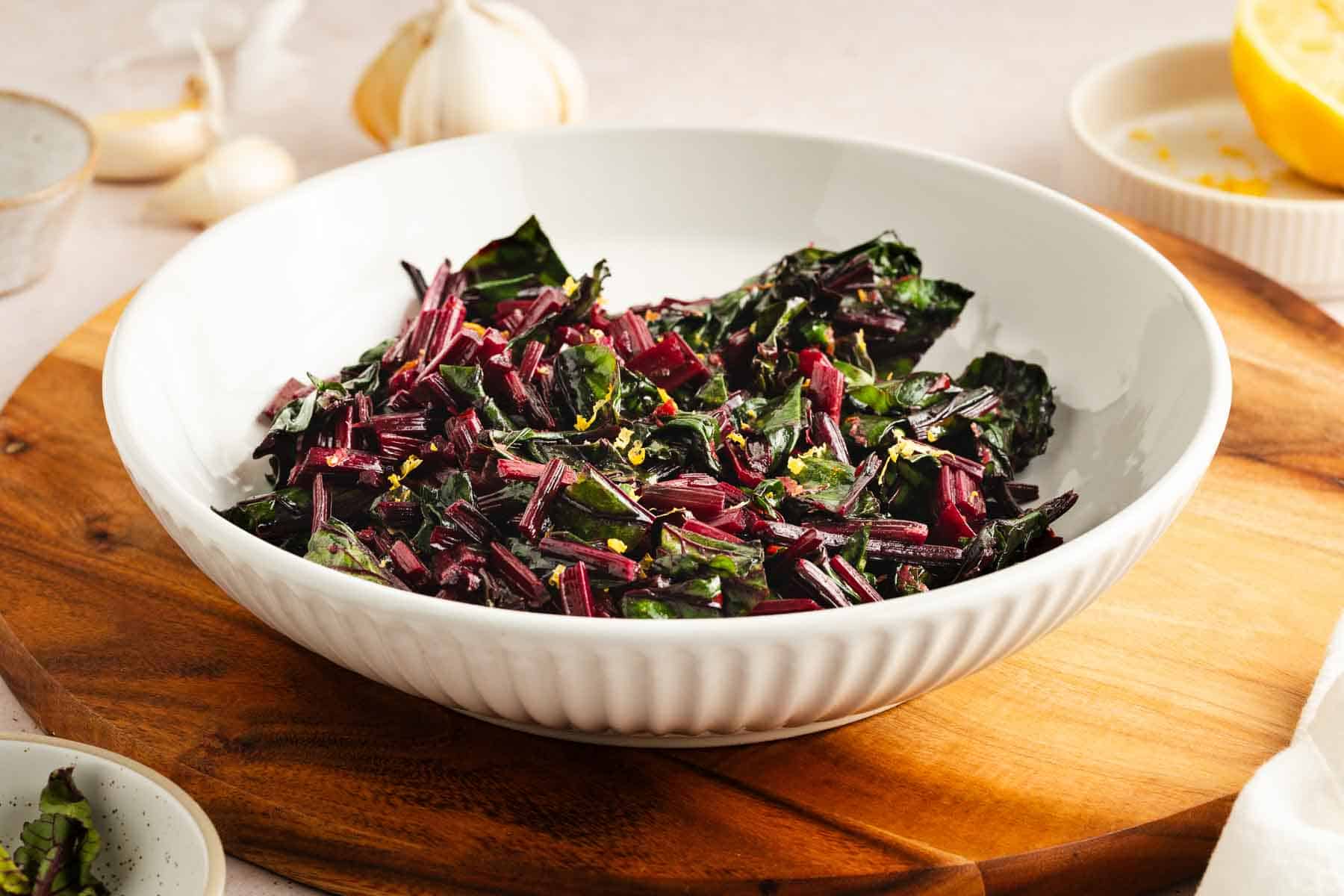 A white bowl filled with sautéed beet greens and stems, garnished with lemon zest, placed on a wooden board with garlic and lemon in the background.
