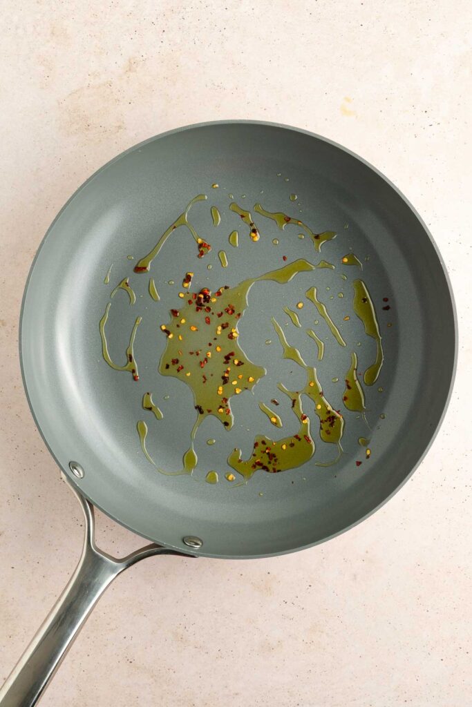 A gray frying pan with a small amount of olive oil and red pepper flakes on its surface, placed on a light-colored countertop.