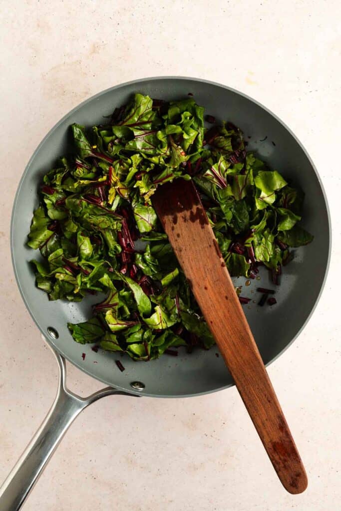 A frying pan with sautéed leafy greens and a wooden spatula resting inside, placed on a light-colored surface.