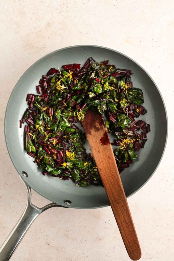 A skillet with sautéed chopped greens and stems, garnished with lemon zest, and a wooden spatula resting inside.