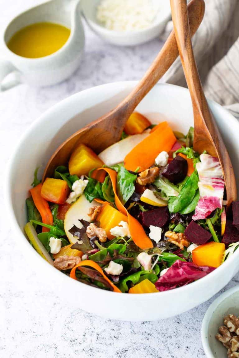 A bowl of mixed salad with leafy greens, sliced carrots, beets, pear, walnuts, and cheese, with wooden salad servers and a small jug of dressing nearby.
