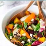 A bowl of roasted beet salad with mixed greens, sliced pears, carrots, walnuts, and crumbled cheese, served with wooden salad utensils.