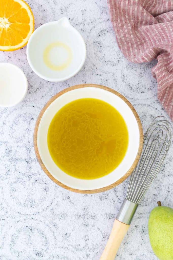 A bowl of yellow liquid, likely dressing or sauce, sits on a light countertop with a whisk, a halved orange, small bowls, and a red cloth nearby.
