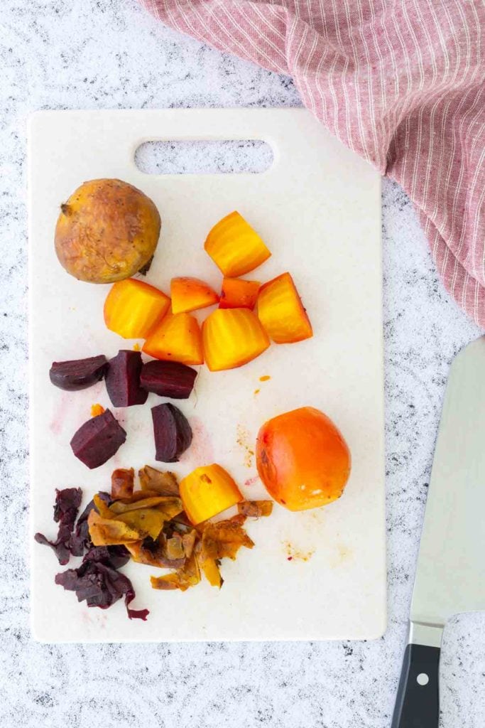A white cutting board with sliced yellow and red beets, beet peelings, a whole yellow beet, a kitchen towel, and a knife.